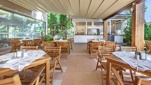 An outdoor dining area with wooden tables and chairs, a kitchen in the background, and a covered patio. - Hotel Sentido Bellevue Beach Sunny Beach (Nessebar)