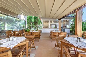 An outdoor dining area with wooden tables and chairs, a kitchen in the background, and a covered patio. - Hotel Sentido Bellevue Beach Sunny Beach (Nessebar)