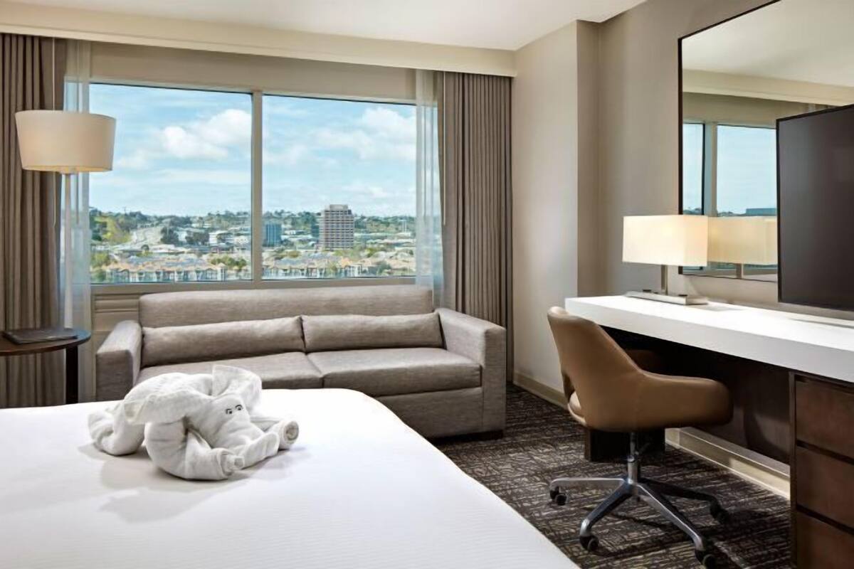 Une chambre d’hôtel comprenant un lit, un canapé, un bureau avec une chaise, une lampe et une grande fenêtre donnant sur des immeubles.