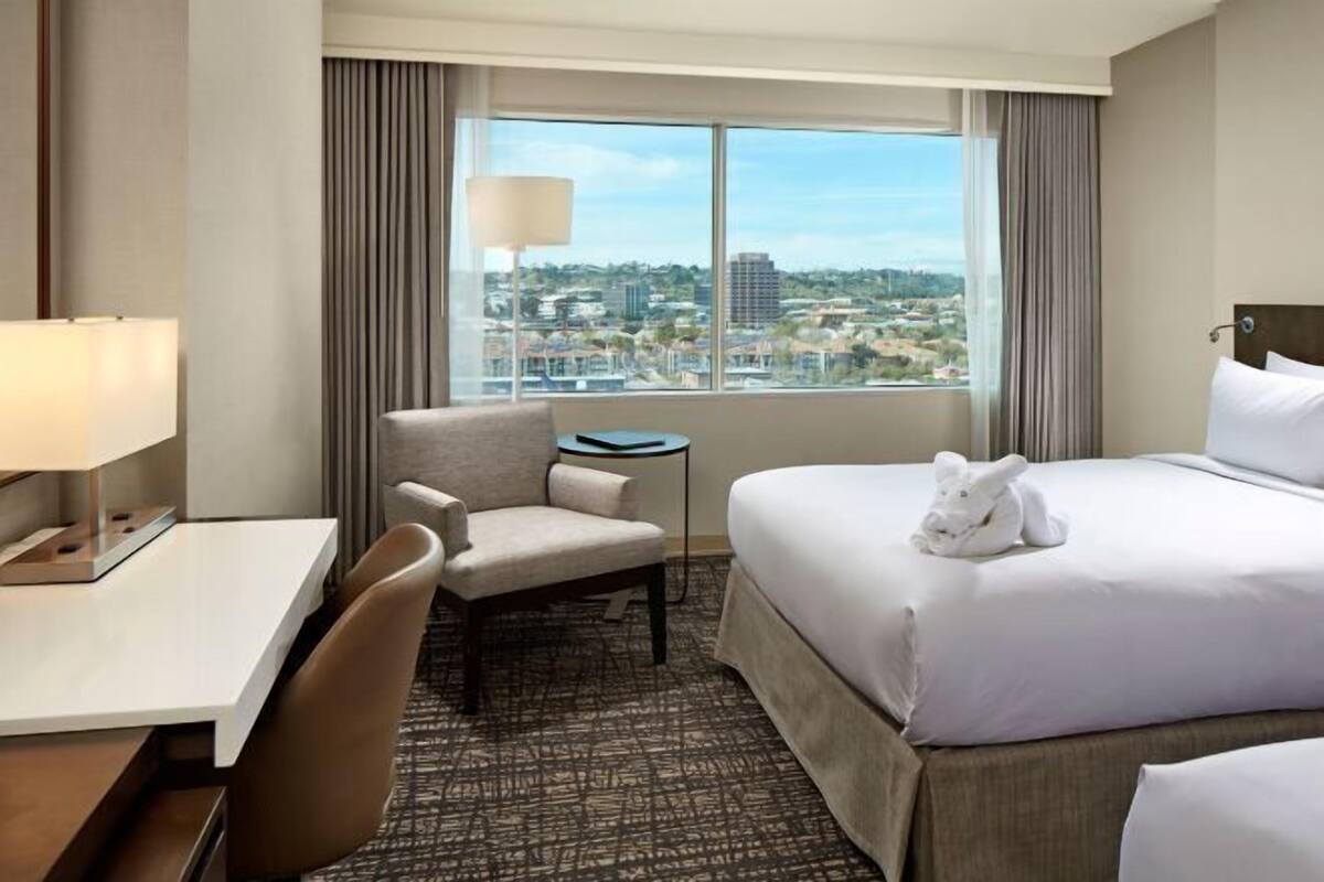 Une chambre d’hôtel équipée d’un lit, d’un bureau, d’une chaise, d’une lampe et offrant une vue sur le paysage urbain grâce à de grandes fenêtres.