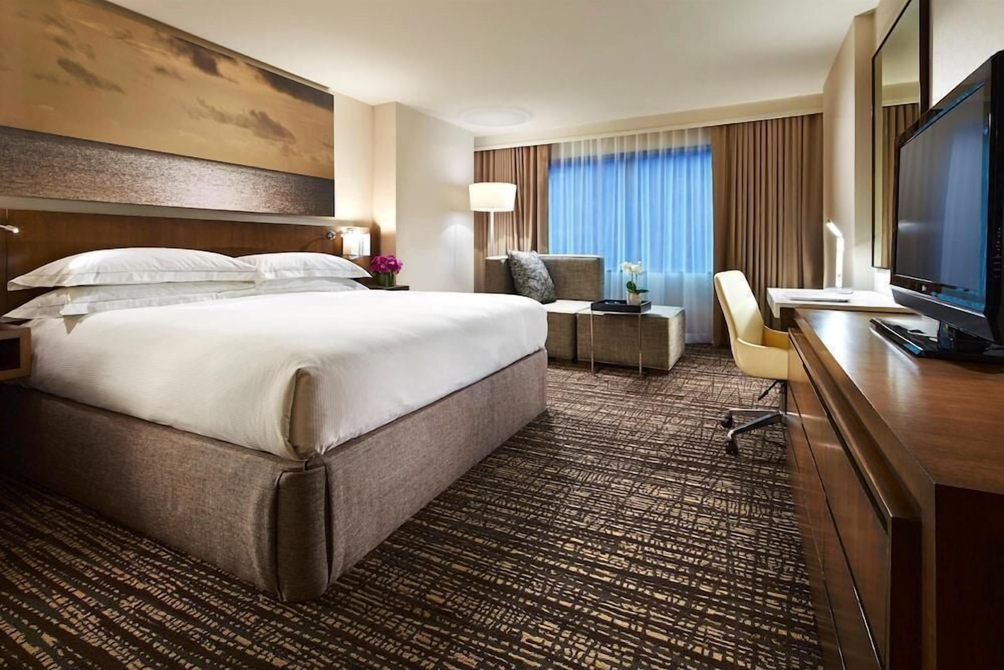 Ein Hotelzimmer mit einem großen Bett, einem Schreibtisch mit Computer, einem Fernseher und einem Fenster mit Vorhängen.