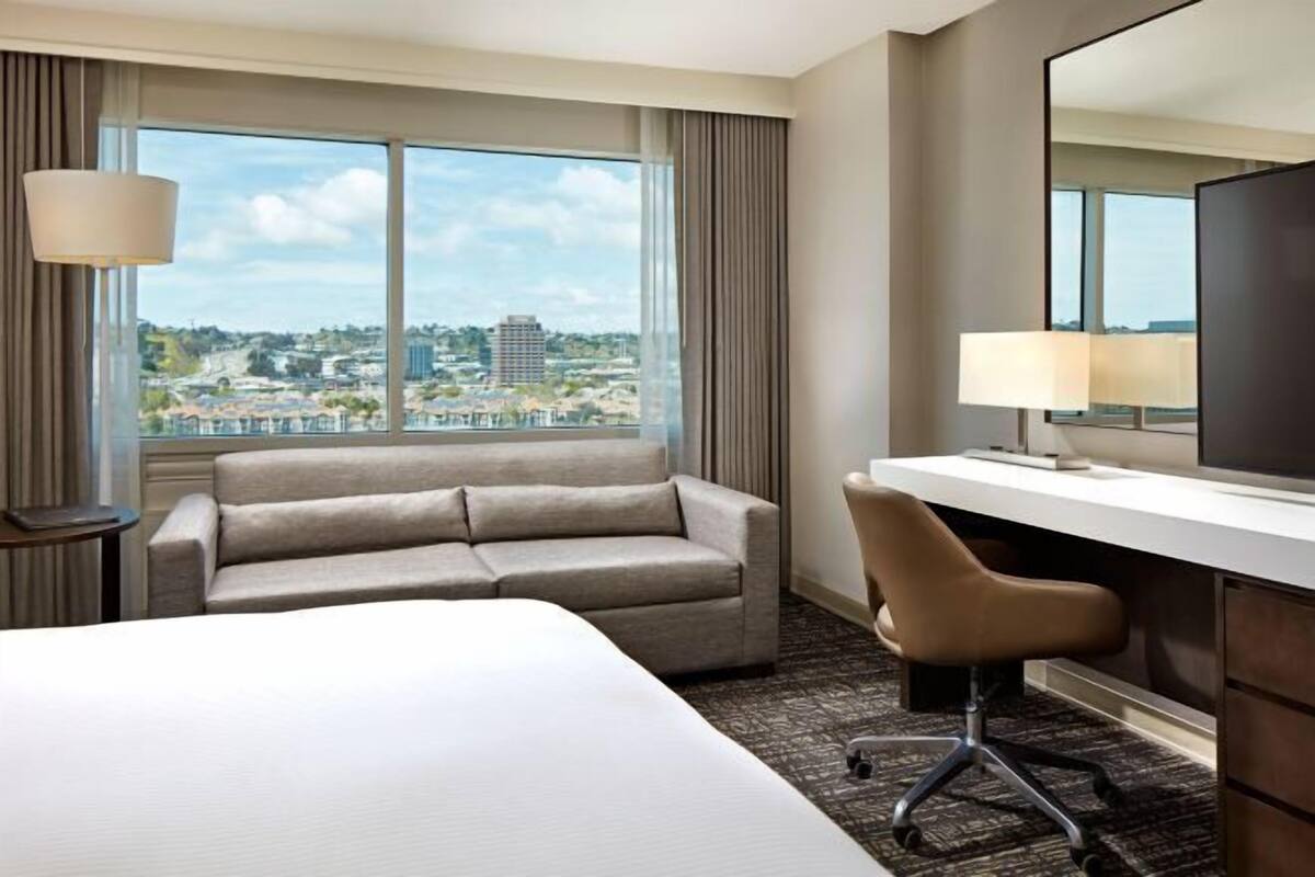 Une chambre d’hôtel comprenant un lit, un canapé, un bureau, une chaise, une lampe et une grande fenêtre donnant sur la ville.