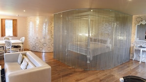 Een kamer met een gebogen glazen scheidingswand, een bed, een witte bank en een eethoek met stoelen en een tafel.