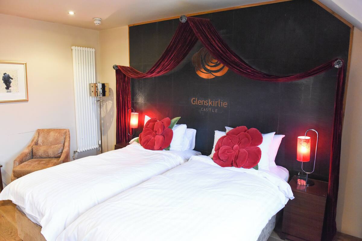 Une chambre d’hôtel avec un grand lit, deux oreillers rouges en forme de fleur, une chaise en bois, un radiateur et un luminaire fixé au mur.