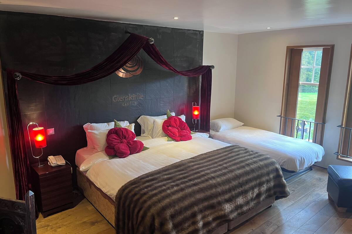 Une chambre avec un grand lit, deux tables de chevet, une commode, une lampe rouge et une fenêtre donnant sur un paysage verdoyant.
