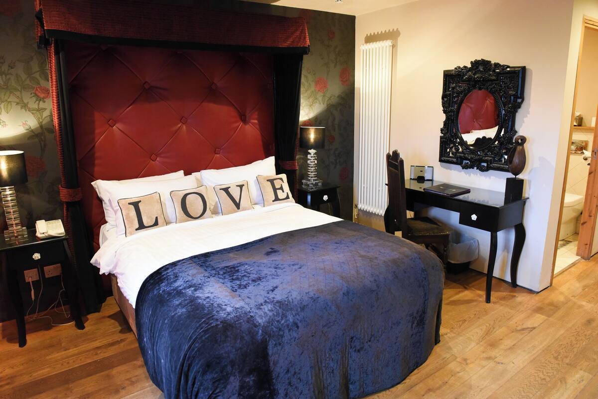 Une chambre à coucher avec une tête de lit capitonnée, un lit agrémenté d’oreillers ornés du mot « AMOUR », un bureau avec un miroir et une lampe.