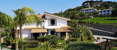 A two-story house with a balcony, surrounded by lush greenery and a stone wall.