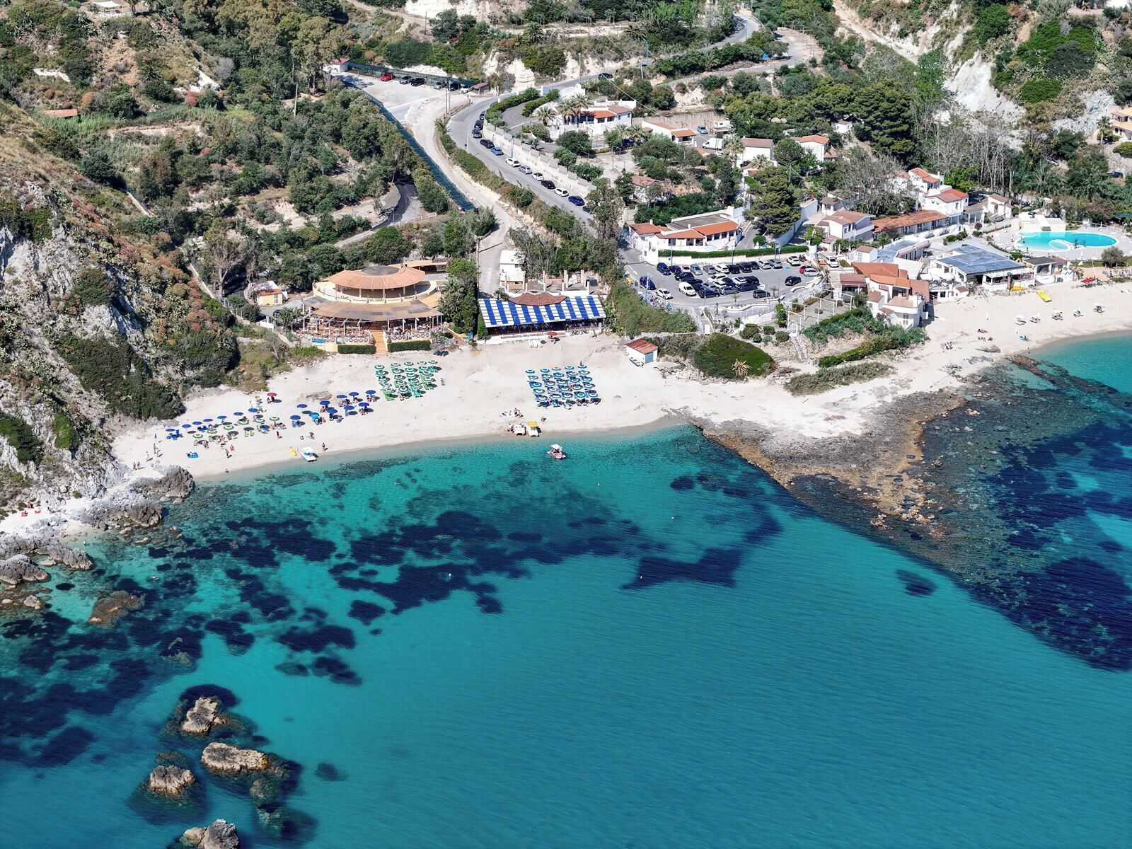 An aerial view of a coastal resort with a sandy beach, clear blue waters, and a variety of buildings including a hotel with a pool and beach umbrellas.