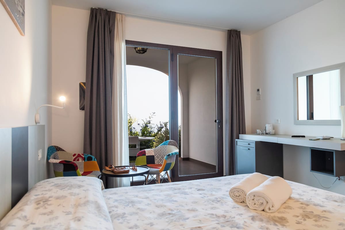 Ett hotellrum med en säng, ett skrivbord, en stol, ett litet bord, en balkong med utsikt och ett badrum.