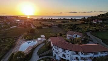 Un edificio a più piani con piscina, immerso nel verde e con vista sul mare al tramonto.