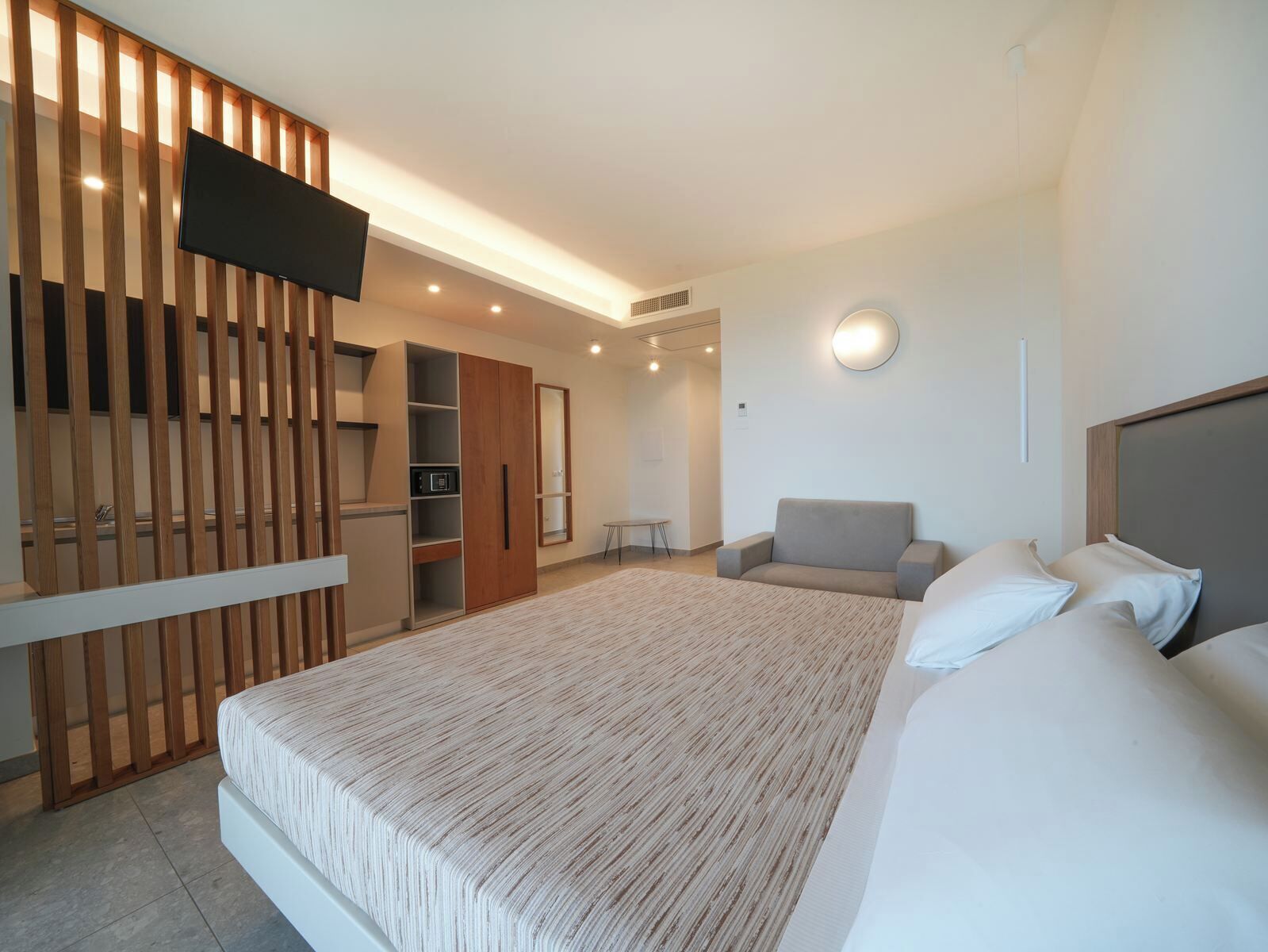 A modern hotel room with a large bed, a grey armchair, a wooden wardrobe, and a flat-screen TV mounted on the wall.