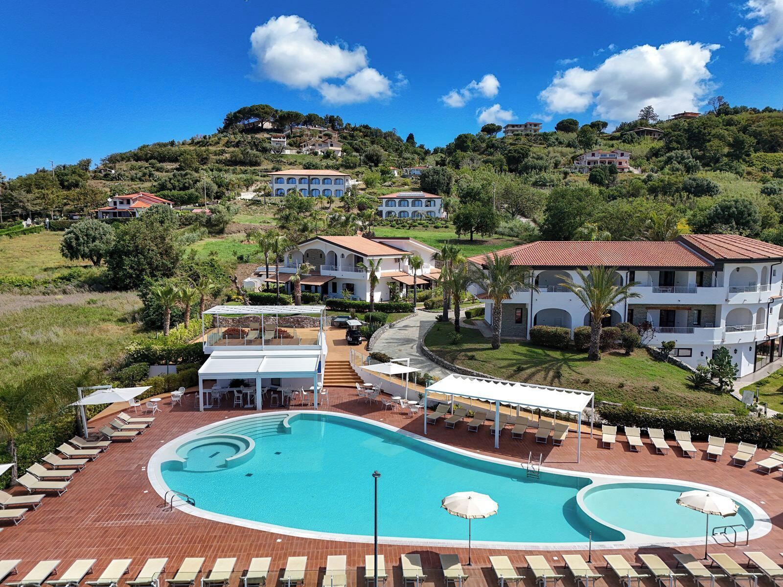 A resort with a large swimming pool, sun loungers, and a view of hillside villas.