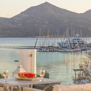 Un puerto deportivo con varios veleros amarrados, una montaña al fondo y una mesa con una copa de vino y una tajada de sandía.