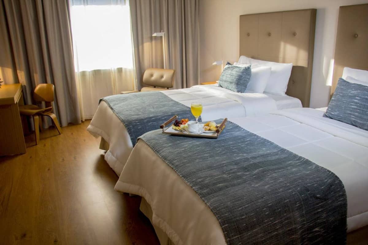 A hotel room with two beds, a desk, and a chair. There is a tray with fruits and a drink on the bed.
