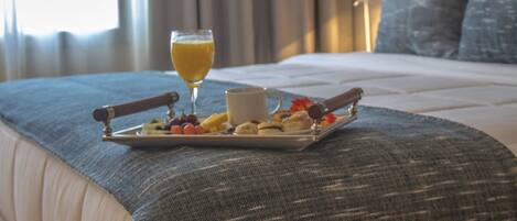 Una cama bien hecha con una bandeja de desayuno que incluye frutas, pasteles y un vaso de zumo de naranja, sobre una colcha con textura.
