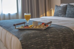 Una cama bien hecha con una bandeja de desayuno que incluye frutas, pasteles y un vaso de zumo de naranja, sobre una colcha con textura.