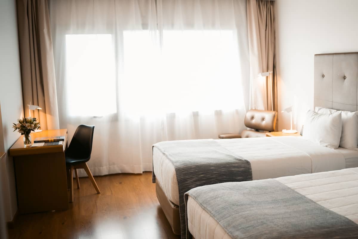 A hotel room with two beds, a desk, a chair, a lamp, and a window with curtains.