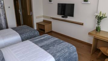A hotel room with two beds, a TV mounted on the wall, a small wooden desk, and a potted plant.