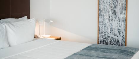 A neatly made bed with white linens and a blue blanket, a wooden headboard, a bedside table with a lamp, and a wall with a tree-like abstract art piece.