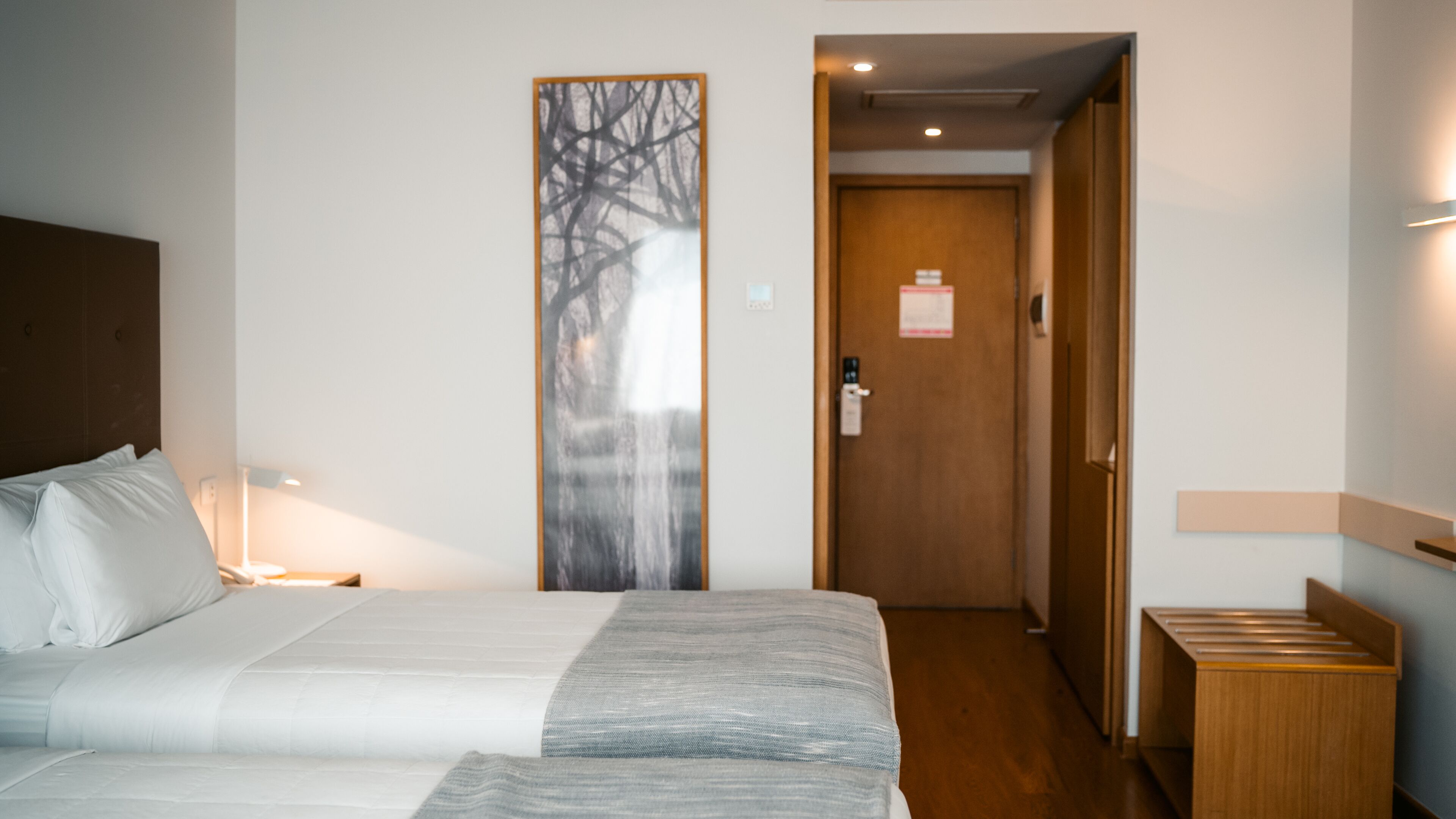 A hotel room with a bed, a nightstand, a wall-mounted lamp, a door with a wooden panel, and a vertical artwork.