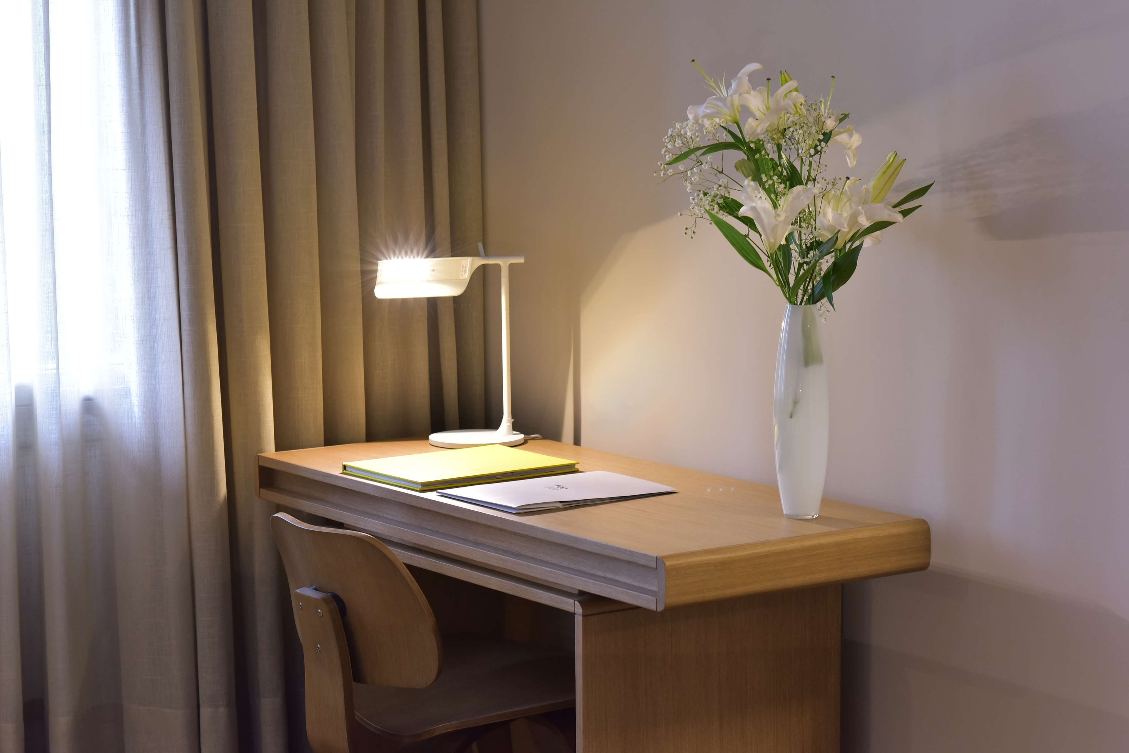 A desk with a lamp, a vase with flowers, and a chair.