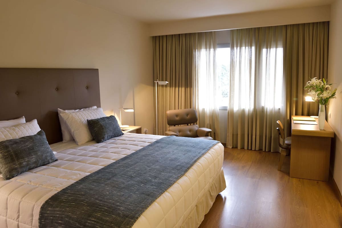 A hotel room with a large bed, a chair, a desk, and a window with curtains.