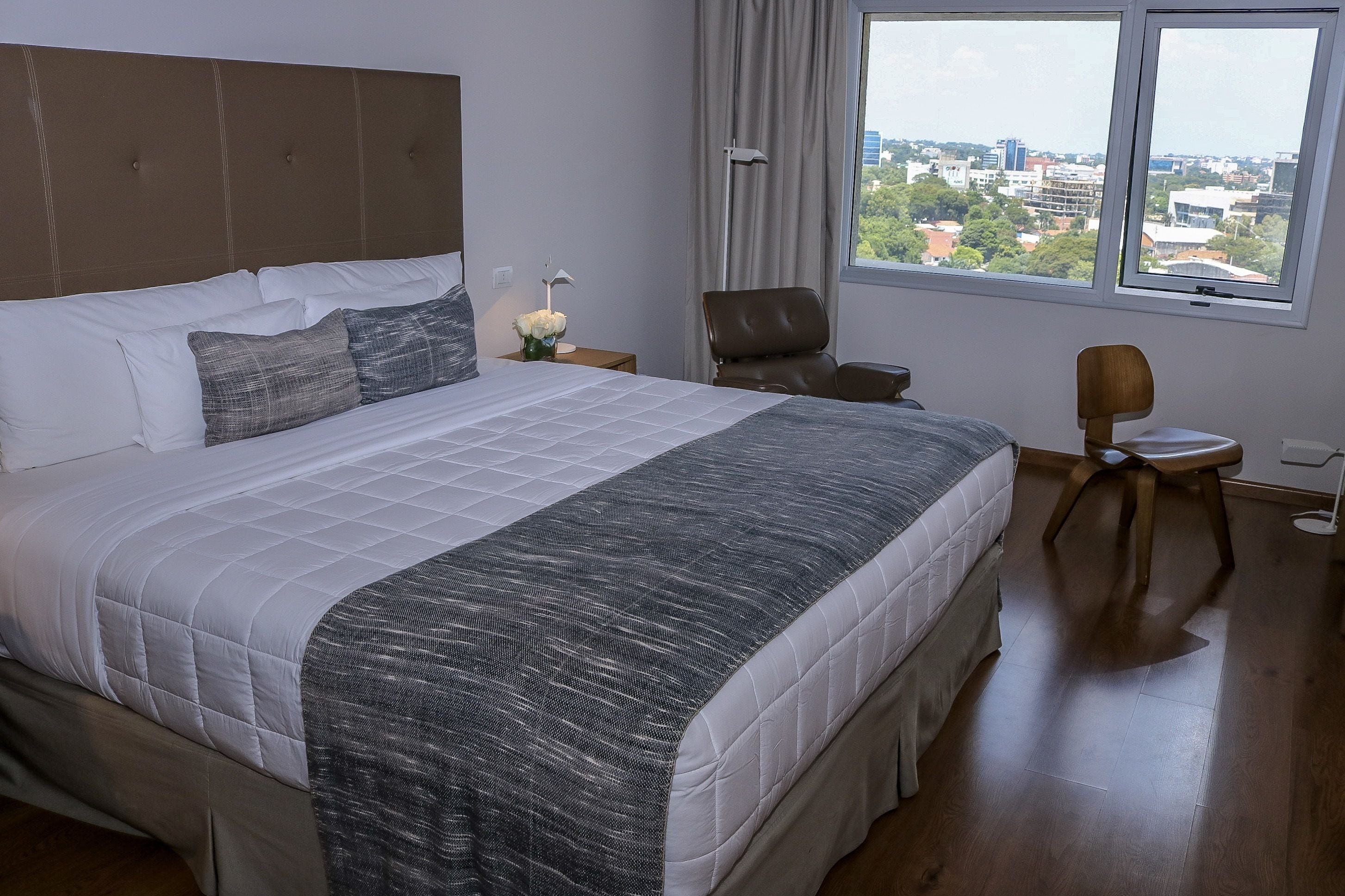 A hotel room with a large bed, a chair, a small table, and a view of the city through a window.