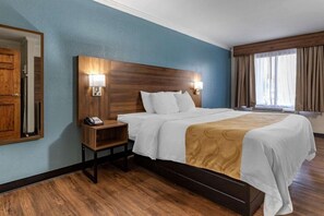 A hotel room with a wooden headboard, a bedside table with a phone, a bed with white bedding and a brown throw, a large window with curtains, and a mirror on the wall. - Quality Inn and Suites Ft Jackson Maingate (Columbia)