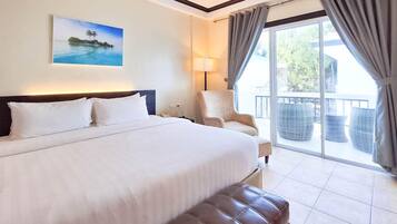 Habitación de hotel con una cama grande, una silla, un balcón con muebles de mimbre y un cuadro de una escena tropical.