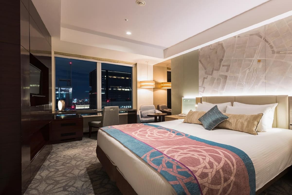 A hotel room with a large bed, a desk, a chair, and a view of a cityscape at night.