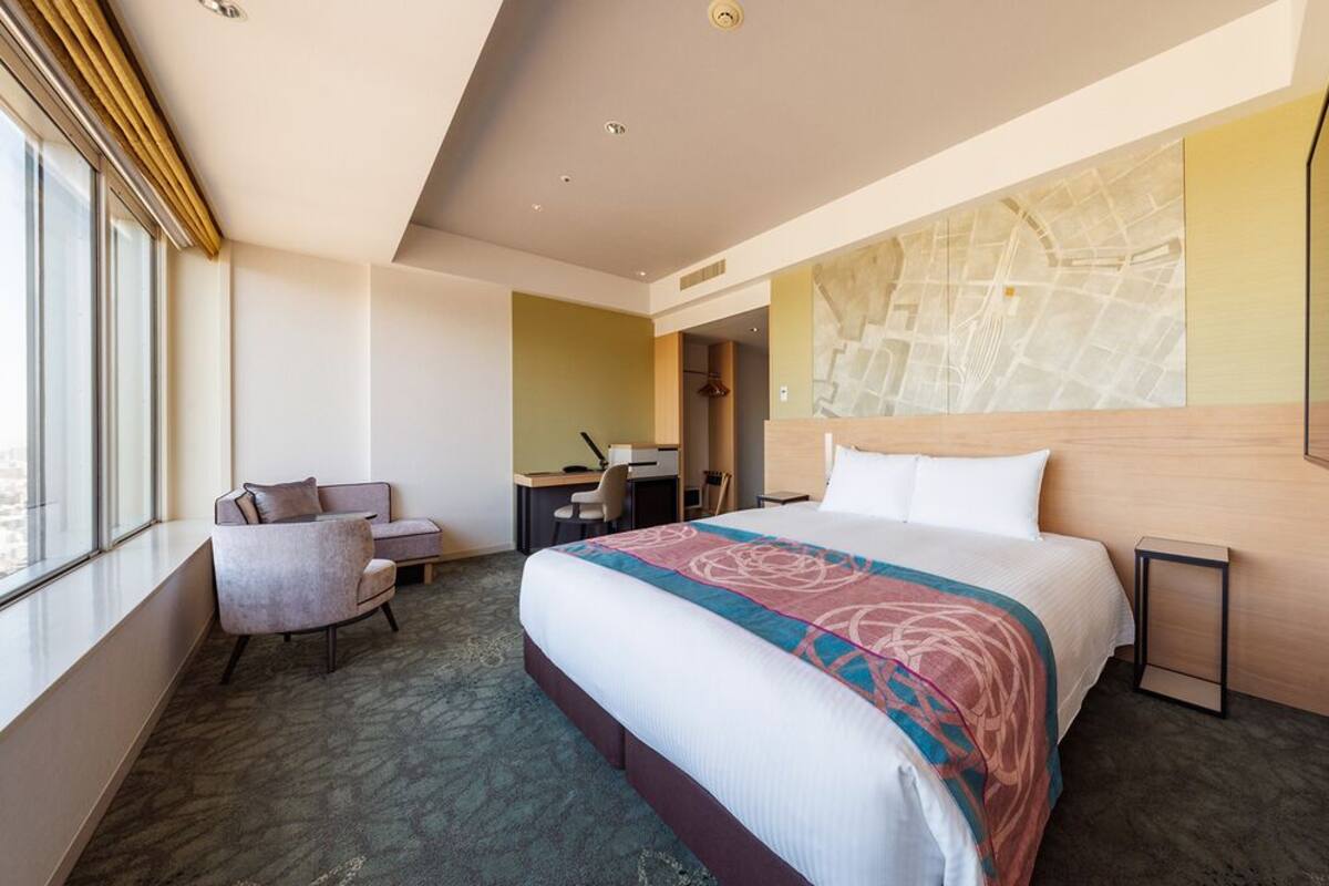 A hotel room with a large bed, a desk, a chair, and a view of the city through large windows.