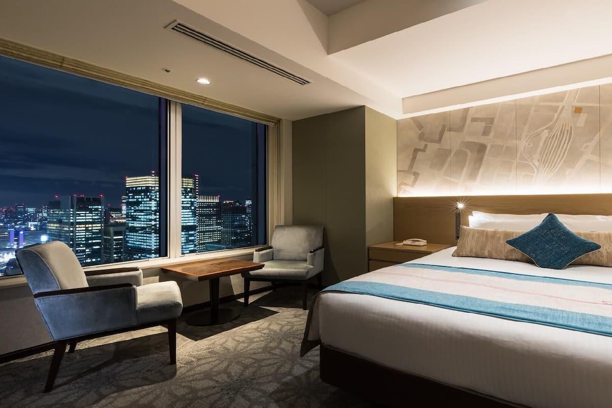 A hotel room with a large bed, a desk, a chair, and a view of a cityscape at night.