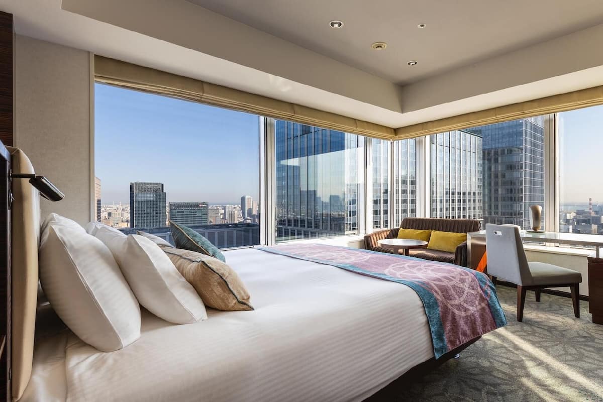 A hotel room with a large bed, a desk, and a chair, with a city view through large windows.