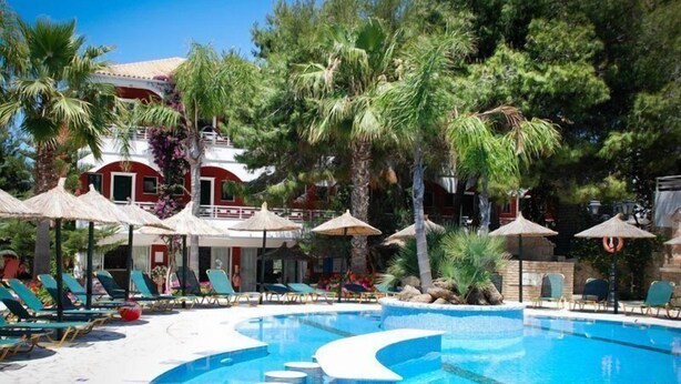 Un área de piscina con sillones de descanso y sombrillas, rodeada de palmeras y un edificio con fachada roja.