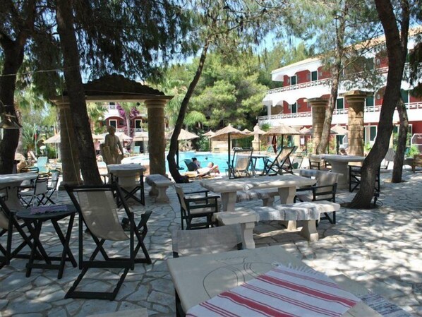 An outdoor seating area with stone tables and chairs, a pool, and a building with red and white walls in the background.