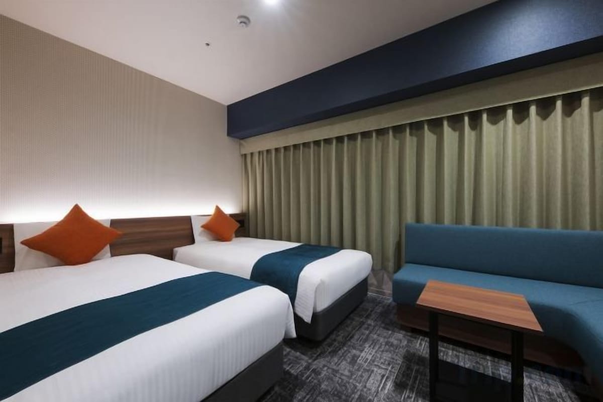 A hotel room with two beds, a sofa, a small table, and a curtain-covered window.