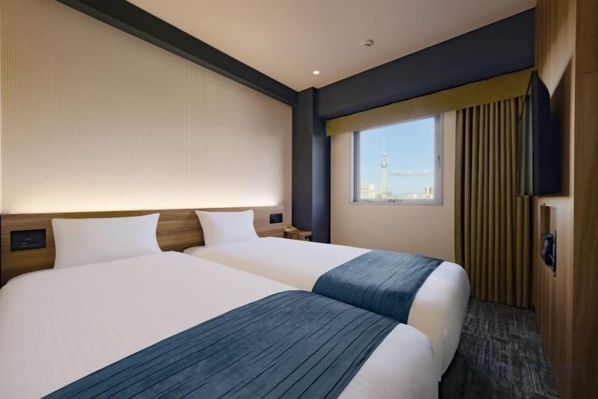 A hotel room with two beds, a large window with a city view, and a flat-screen TV mounted on the wall.