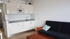 A compact kitchen with white cabinets, a microwave, and a sink. There's a black sofa with a patterned pillow and a wooden table. - Montesol Atsol (Mogan)