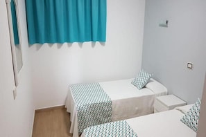 A small, clean room with two single beds, a window with a blue curtain, and a white wall. - Montesol (Mogan)