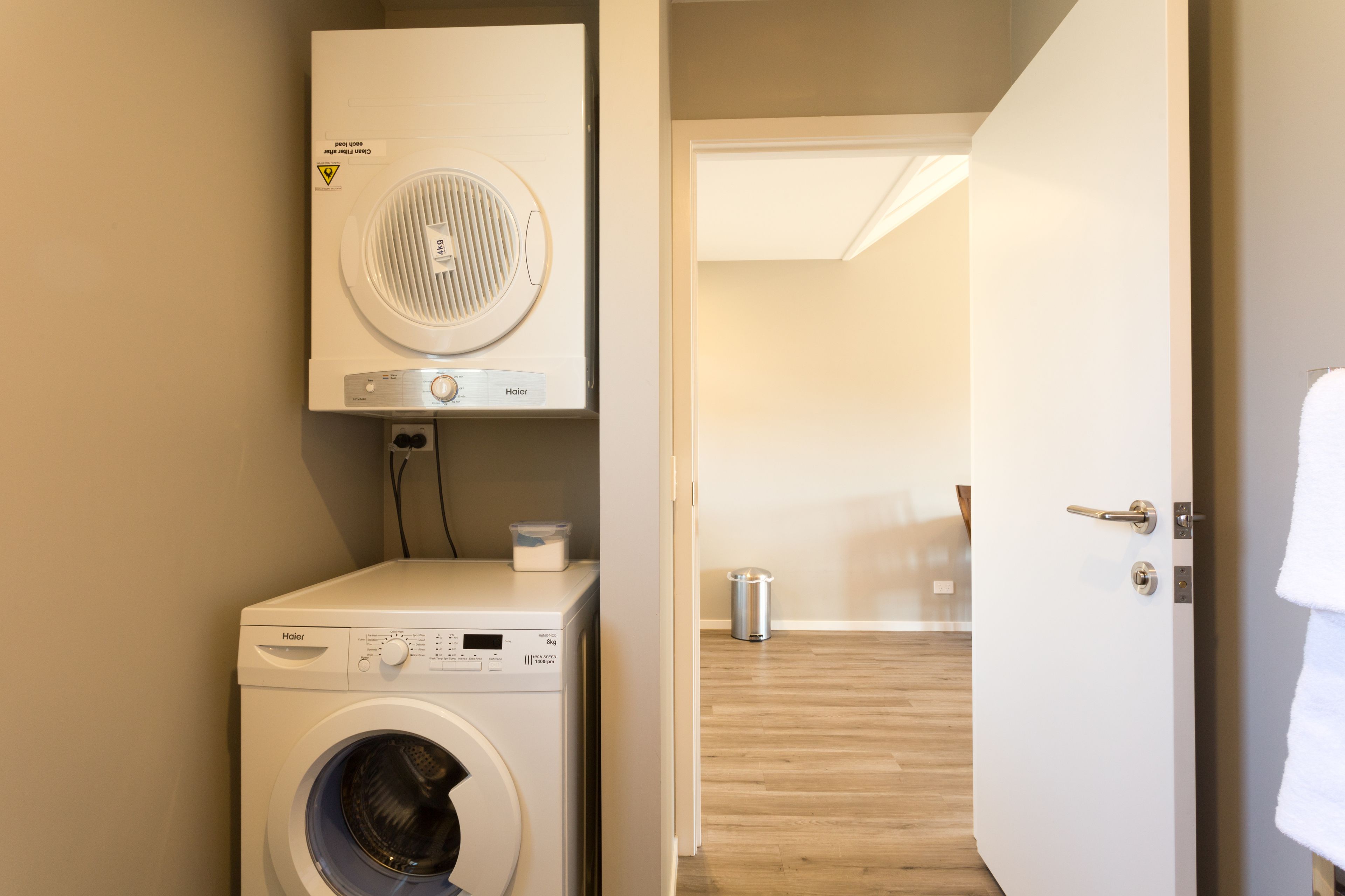 A laundry room with a front-loading washer and dryer, a trash can, and a door leading to another room.