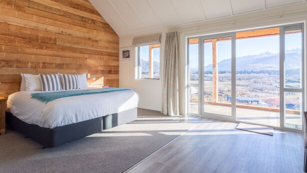 A bedroom with a large bed, wooden wall paneling, a view of mountains, and a sliding glass door. - Shotover Country Cottages (Lower Shotover)