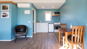 A compact kitchen with white cabinets, a microwave, and a small refrigerator. There is a dining table with chairs and a black leather chair. - Shotover Country Cottages (Lower Shotover)