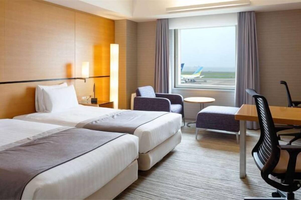 Une chambre d’hôtel avec deux lits, un bureau, une chaise, une petite table et une vue sur l’aéroport.