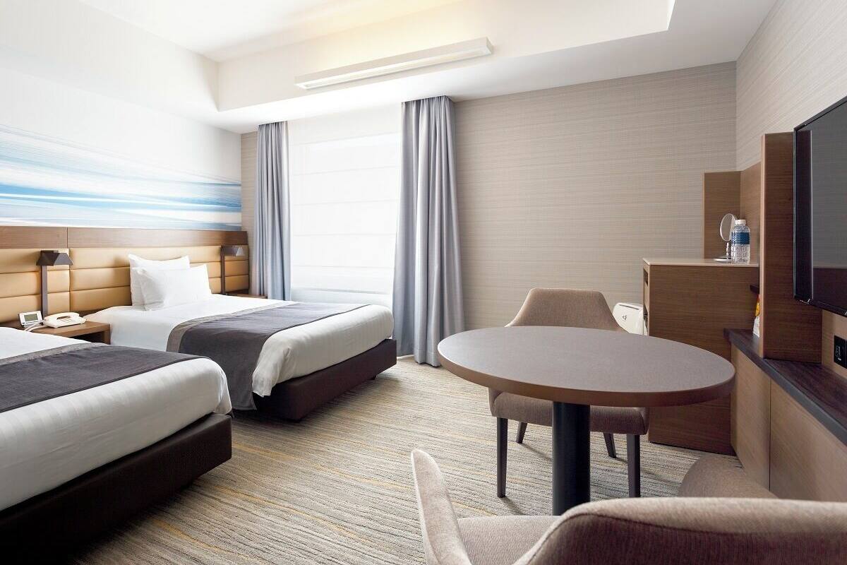 Une chambre d’hôtel avec deux lits, un bureau, une chaise, une télévision et une grande fenêtre avec des rideaux.