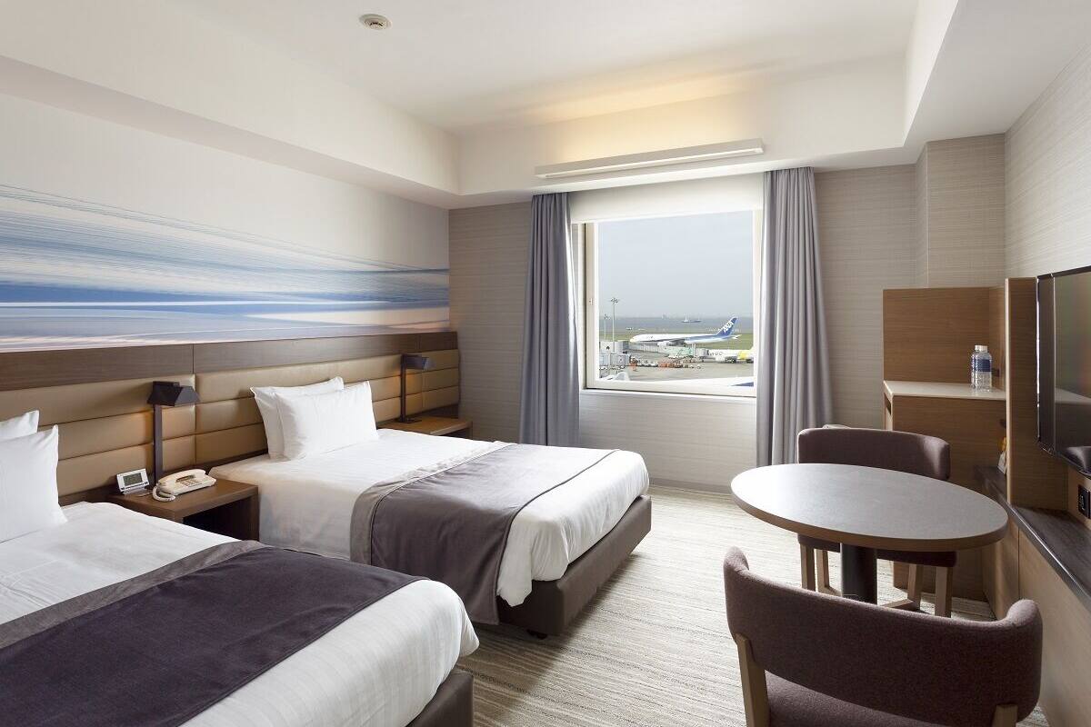 Une chambre d’hôtel avec deux lits, un bureau, une chaise, une télévision et une fenêtre donnant sur l’aéroport.