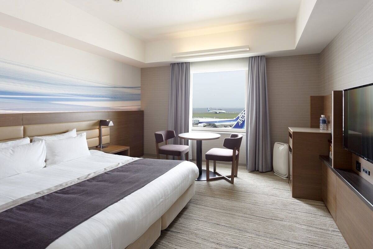 Une chambre d’hôtel avec un lit, un bureau, une télévision, une fenêtre donnant sur un aéroport et un tableau accroché au mur.