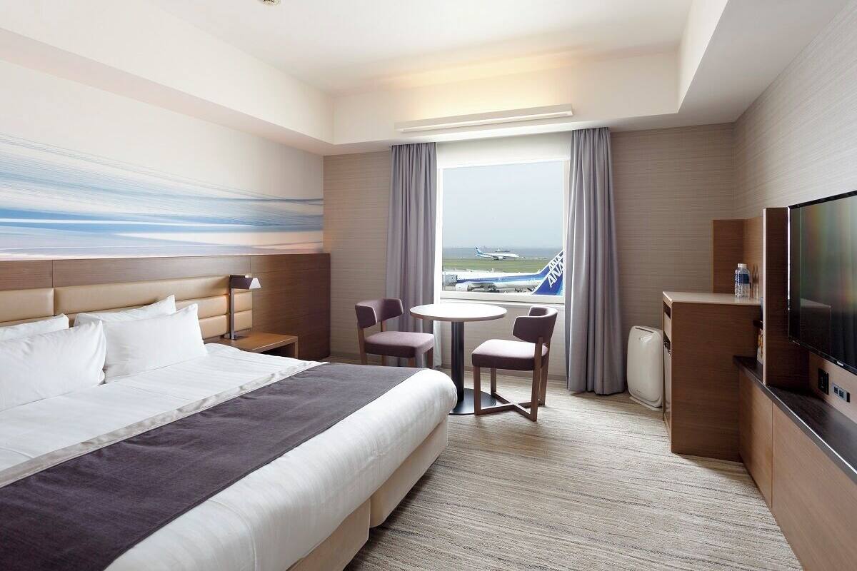 Une chambre d’hôtel avec un lit, un bureau, une télévision, une fenêtre donnant sur un aéroport et un tableau au mur.