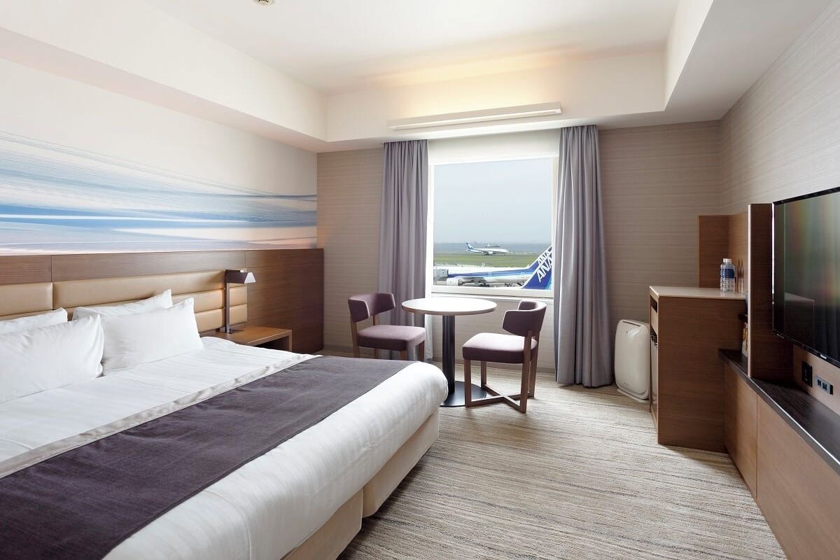 Une chambre d’hôtel avec un lit, un bureau, une télévision, une fenêtre donnant sur un aéroport et un tableau au mur.