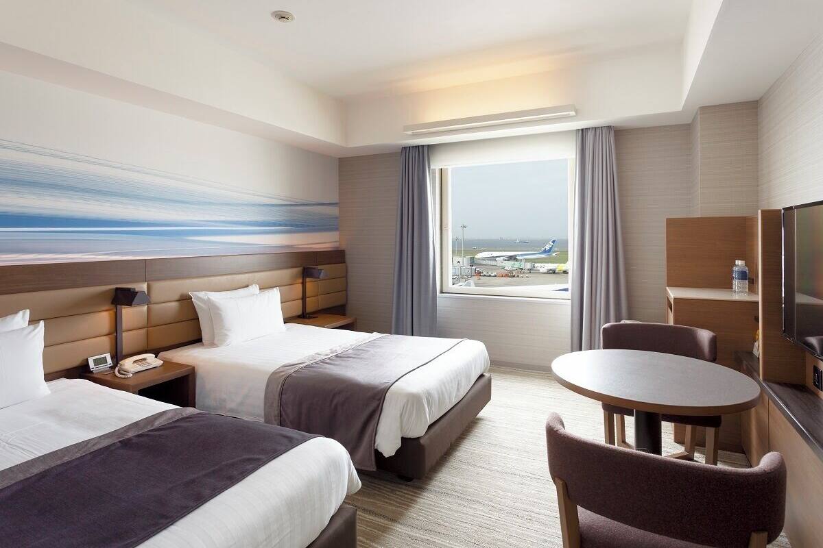 Une chambre d’hôtel avec deux lits, un bureau, une chaise, une télévision et une fenêtre donnant sur l’aéroport.