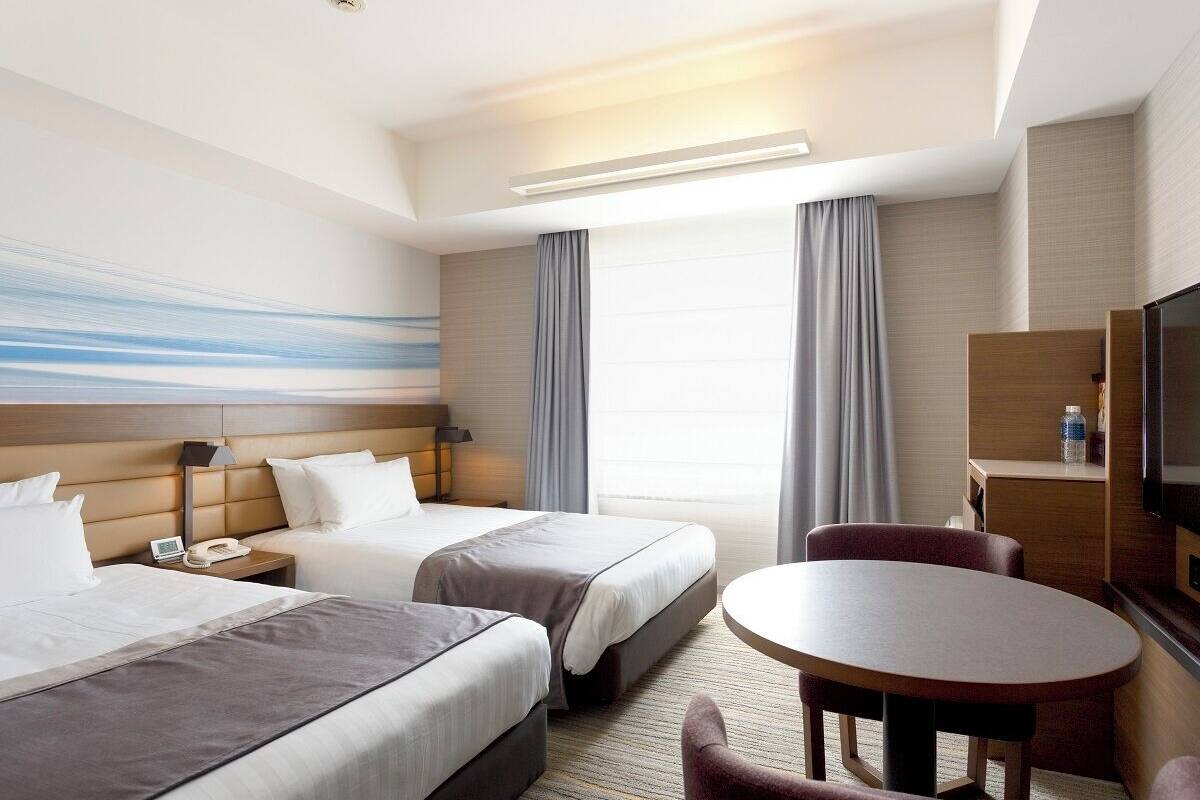 Une chambre d’hôtel avec deux lits, un bureau, une chaise, une télévision et une grande fenêtre avec des rideaux.